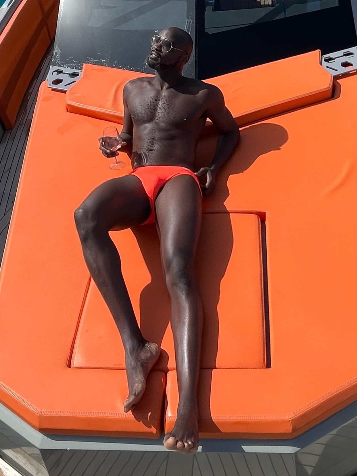 A shirtless man in orange swimwear is sunbathing on an orange lounge chair. He is holding a glass and wearing sunglasses.