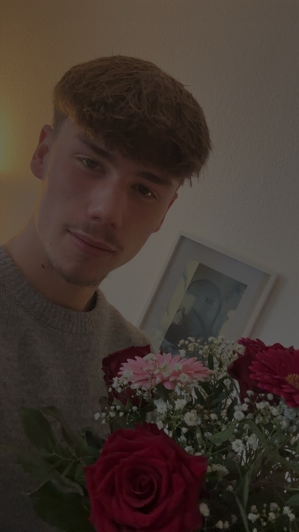 A man holds a bouquet of flowers, looking directly at the camera. The image is a close-up portrait, with a soft, warm lighting.