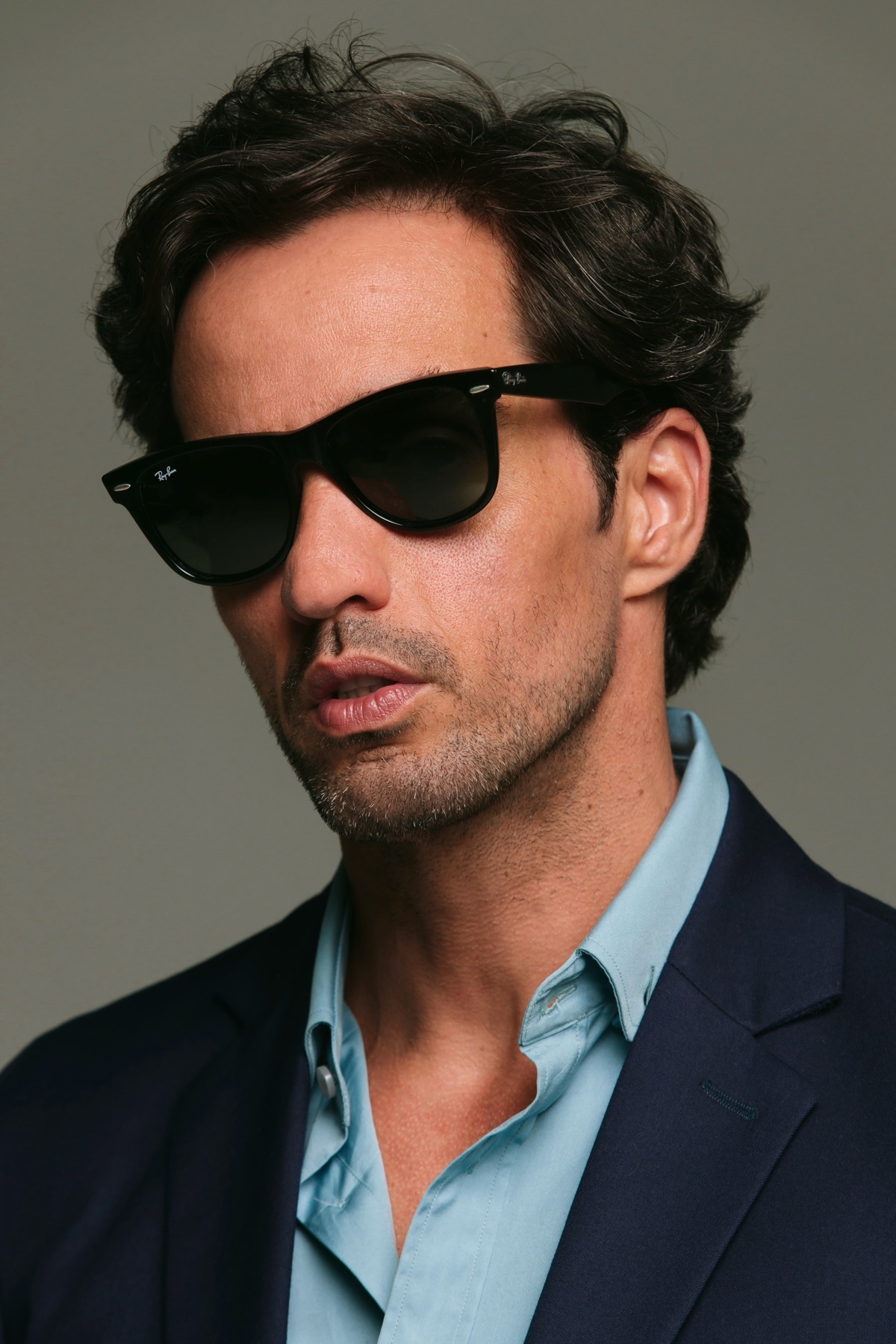 A man wearing sunglasses and a suit poses for a portrait. He has dark, wavy hair and stubble.