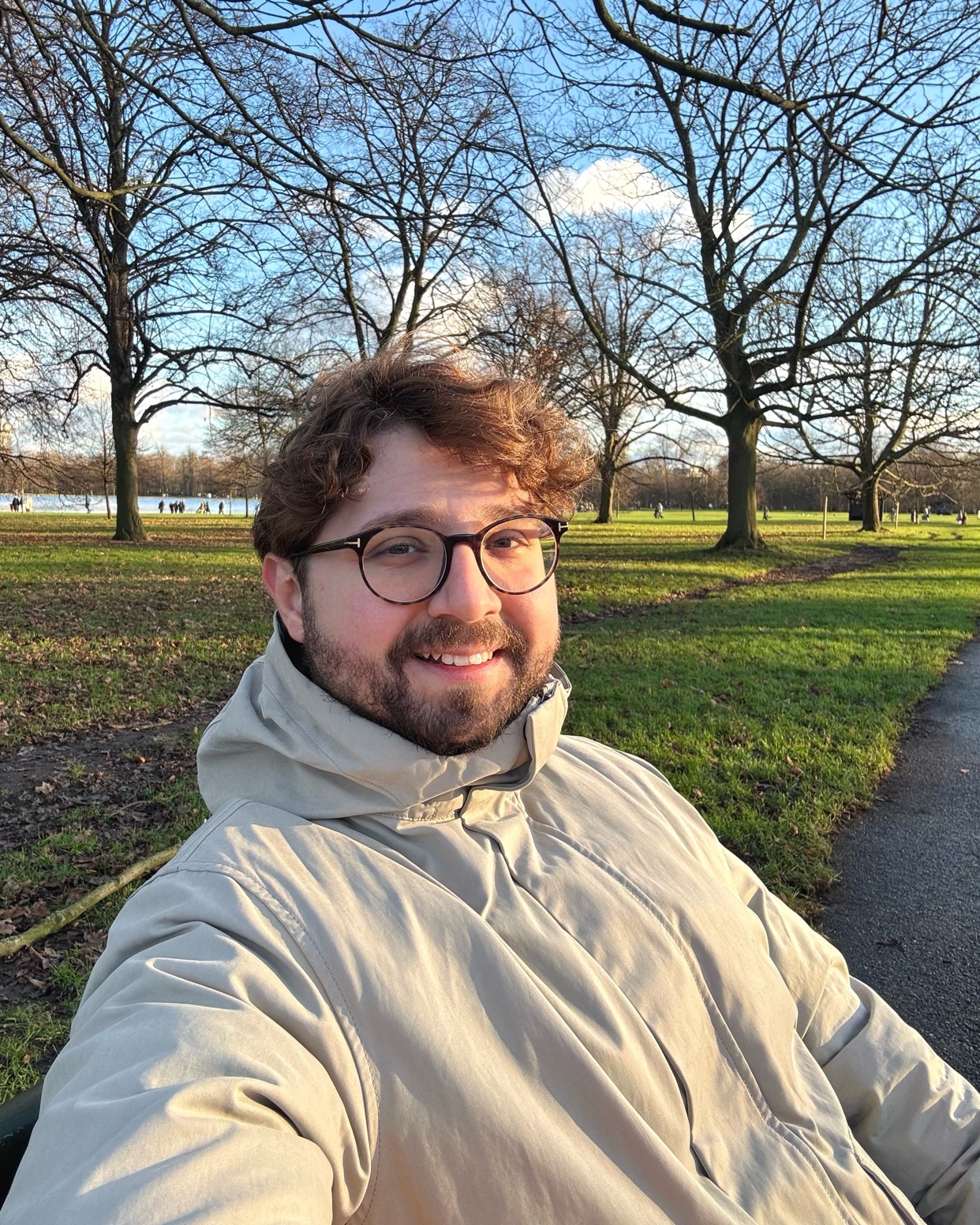 A man smiles for a selfie in a park on a sunny day. He is wearing glasses and a jacket.