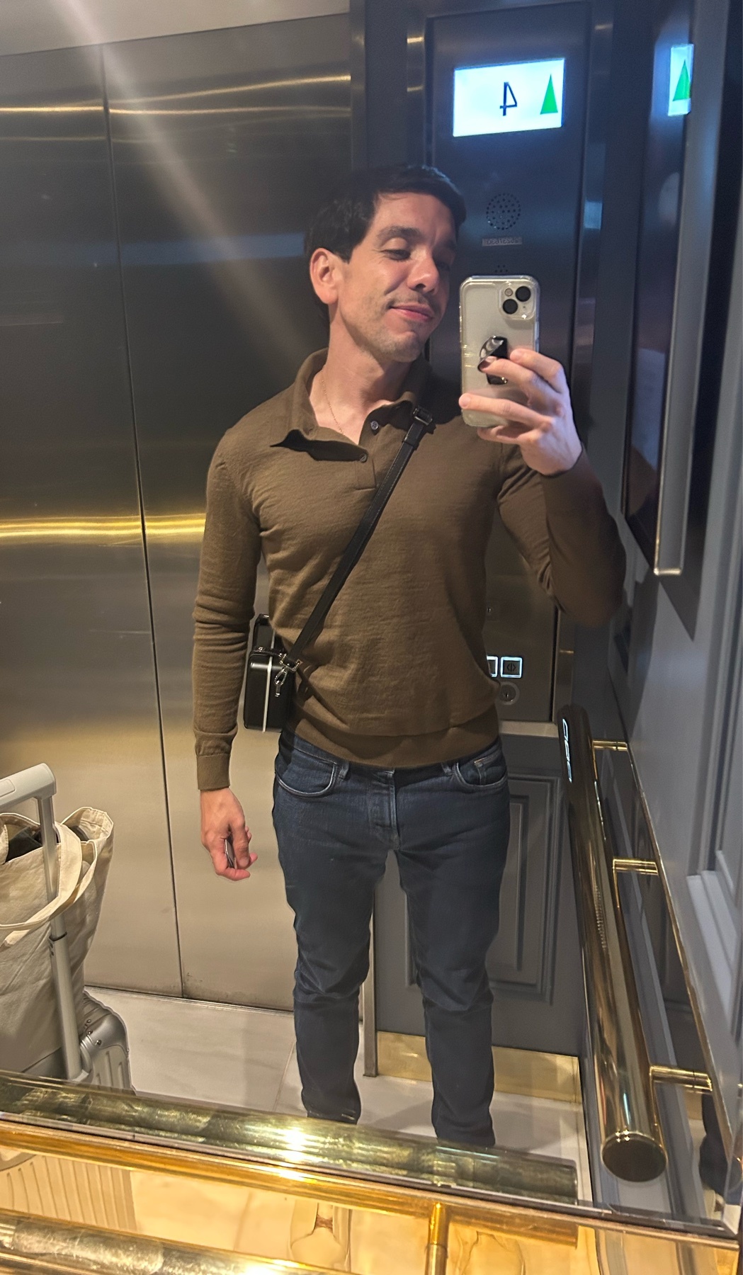 A man takes a selfie in an elevator, wearing a brown polo shirt and jeans.