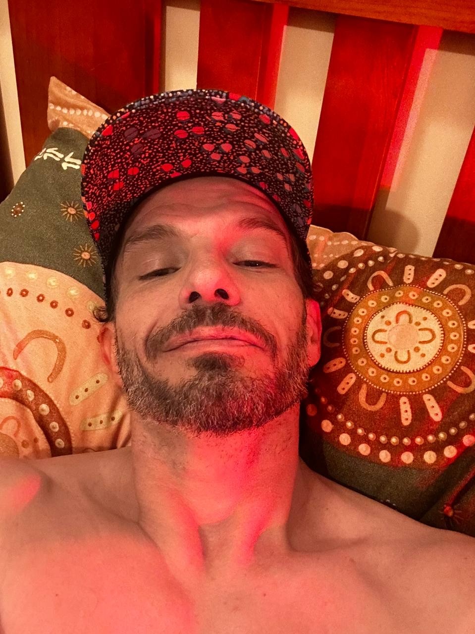 A man wearing a baseball cap and a goatee lies in bed, looking at the camera. The image is illuminated with a red light.