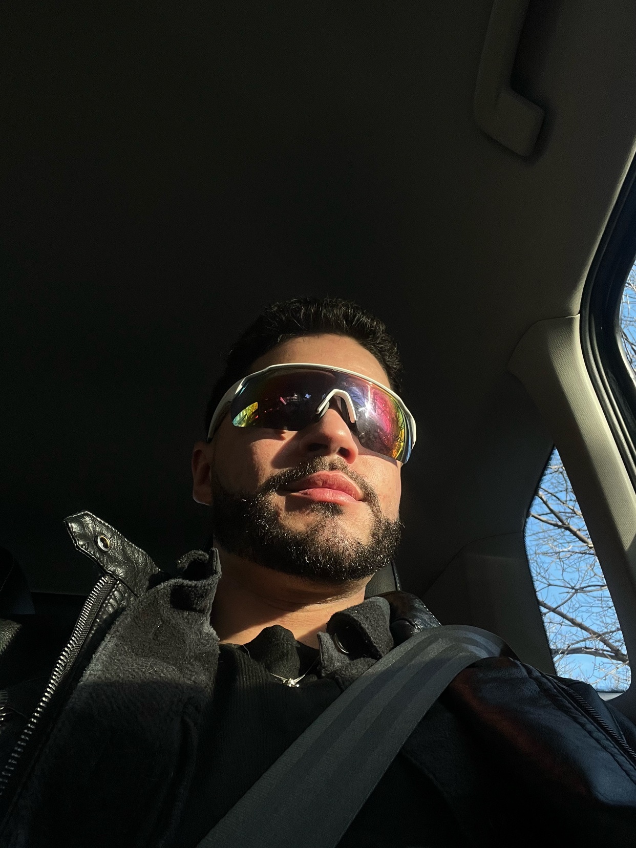 A man wearing sunglasses takes a selfie while driving. The photo is taken from a low angle, with the sun shining through the window.