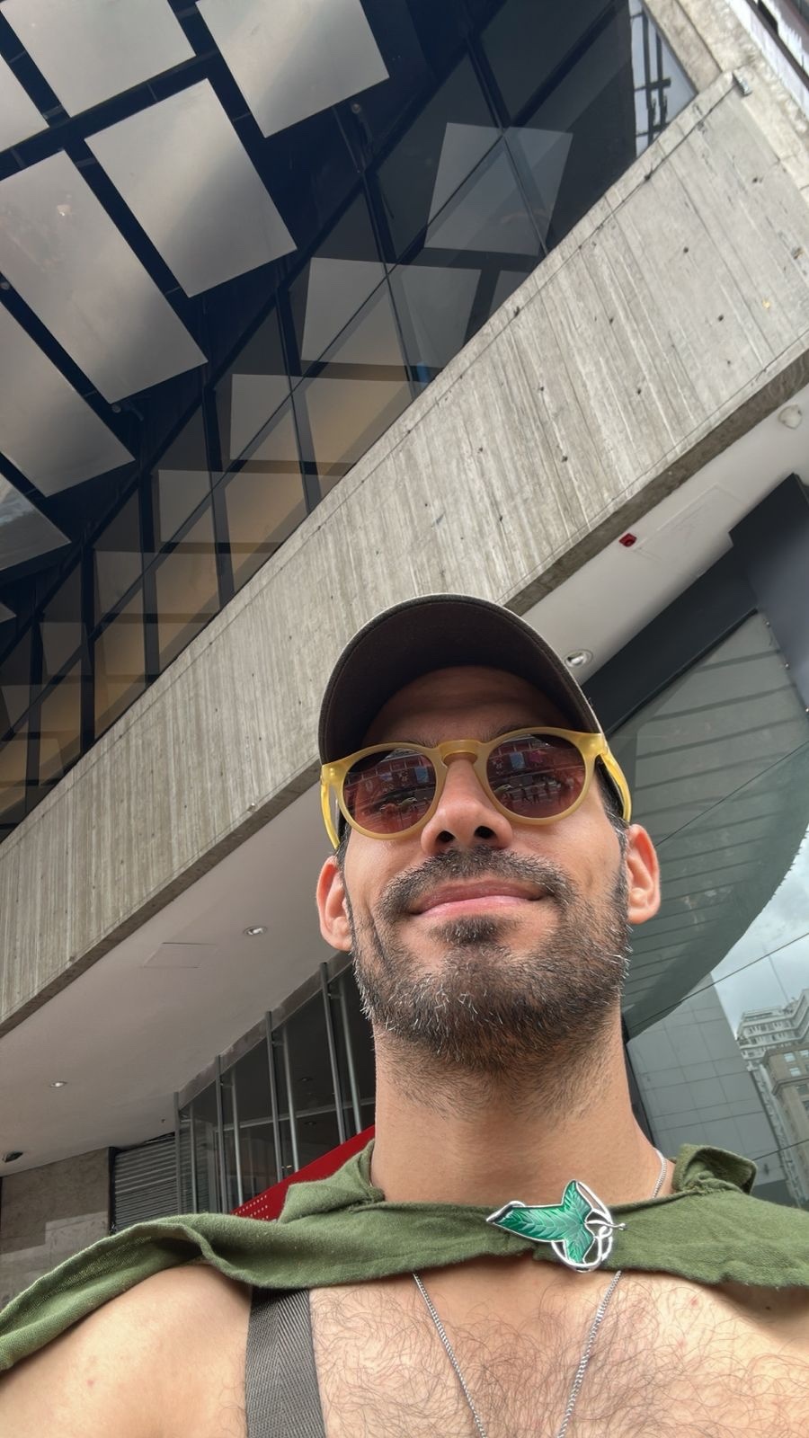 A man wearing sunglasses and a baseball cap takes a selfie in front of a modern building. He is wearing a green cape draped over his shoulders.