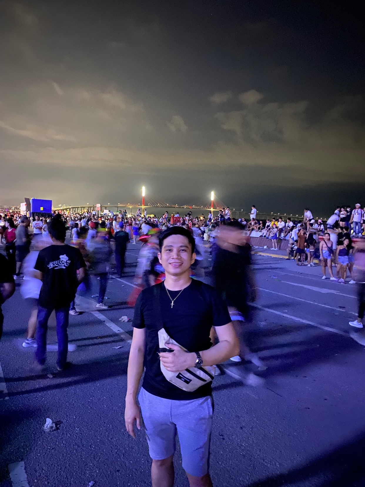 A man smiles at the camera in front of a large crowd at night. He is wearing a black t-shirt, shorts, and a waist bag.
