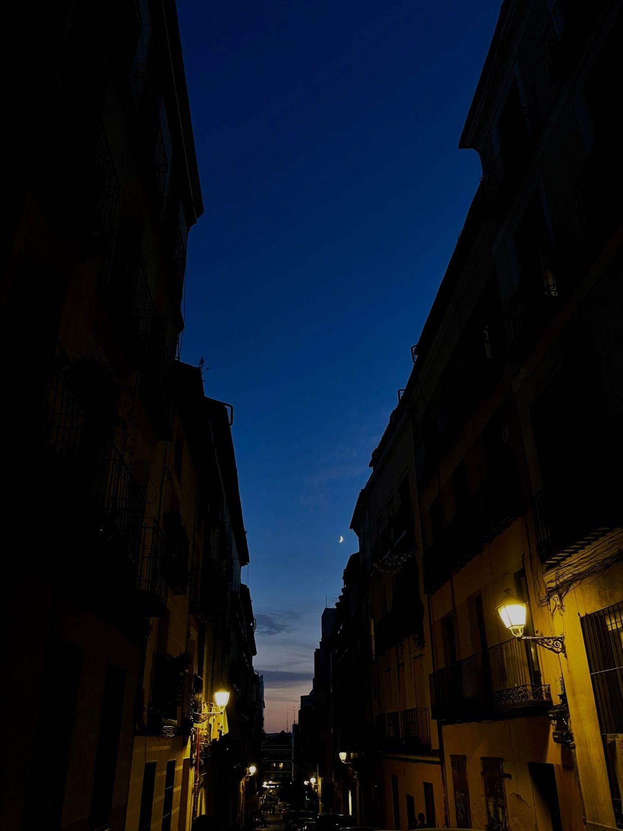 A dark, moody cityscape at night. The image captures a street lined with buildings under a deep blue sky.
