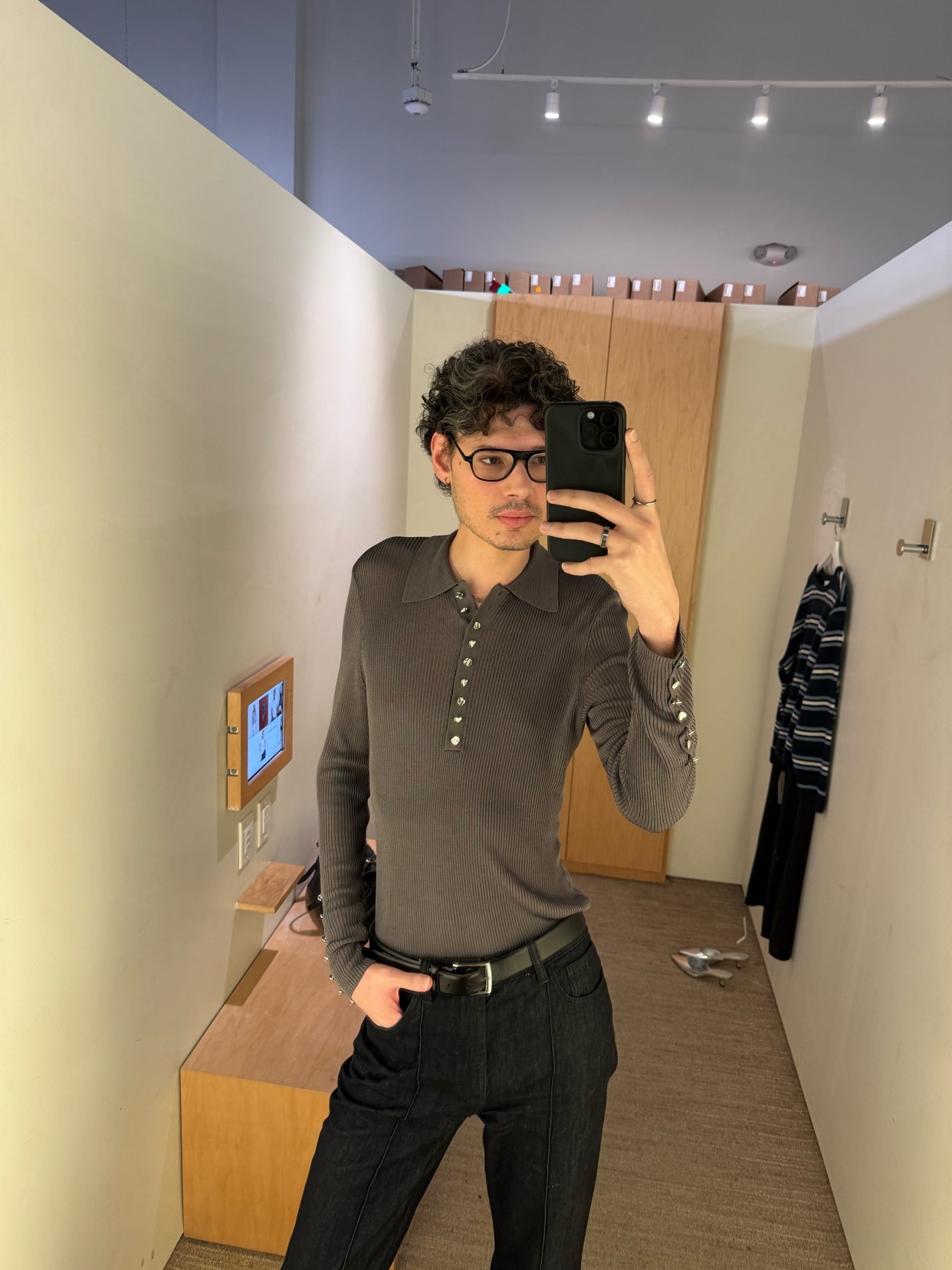 A man takes a mirror selfie in a dressing room, showcasing his outfit. He wears glasses, a long-sleeved shirt, and dark jeans.