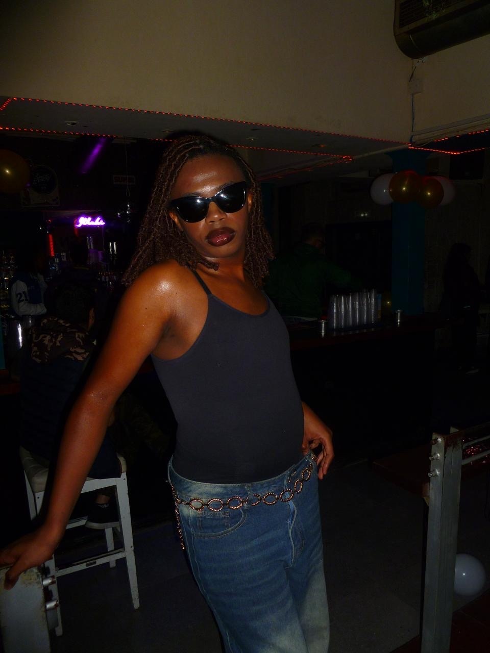 A person in drag poses for a photo. They are wearing sunglasses, a tank top, and jeans.