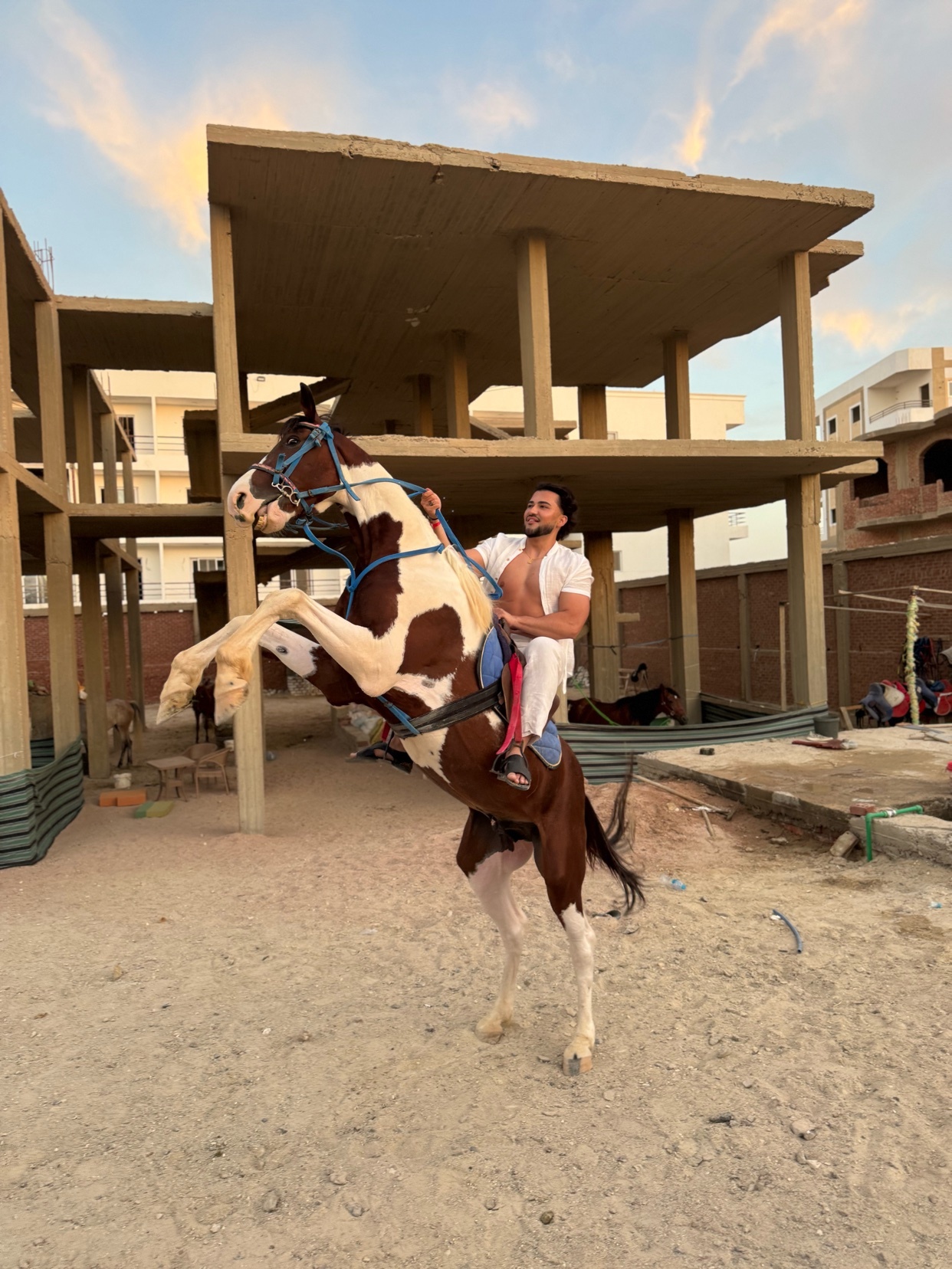 A shirtless man in white pants and a cowboy hat is riding a brown and white horse that is rearing up on its hind legs. The scene appears to be on a construction site with unfinished buildings in the background.