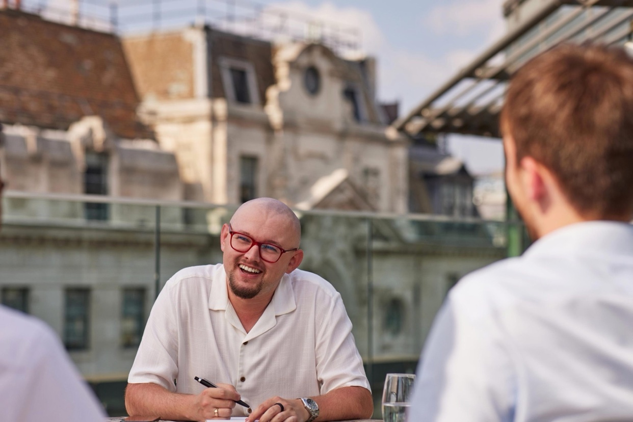 A bald man with glasses smiles while sitting at a table on a rooftop, holding a pen and looking at papers. The background shows a cityscape with old buildings.