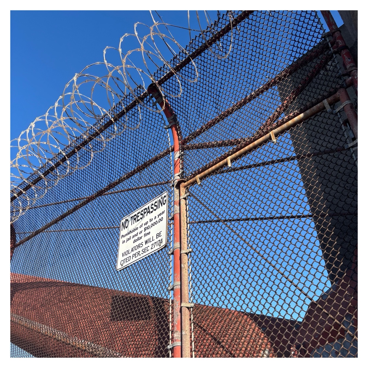 A close-up shot of a fence with barbed wire on top, with a "No Trespassing" sign. The sky is visible in the background.