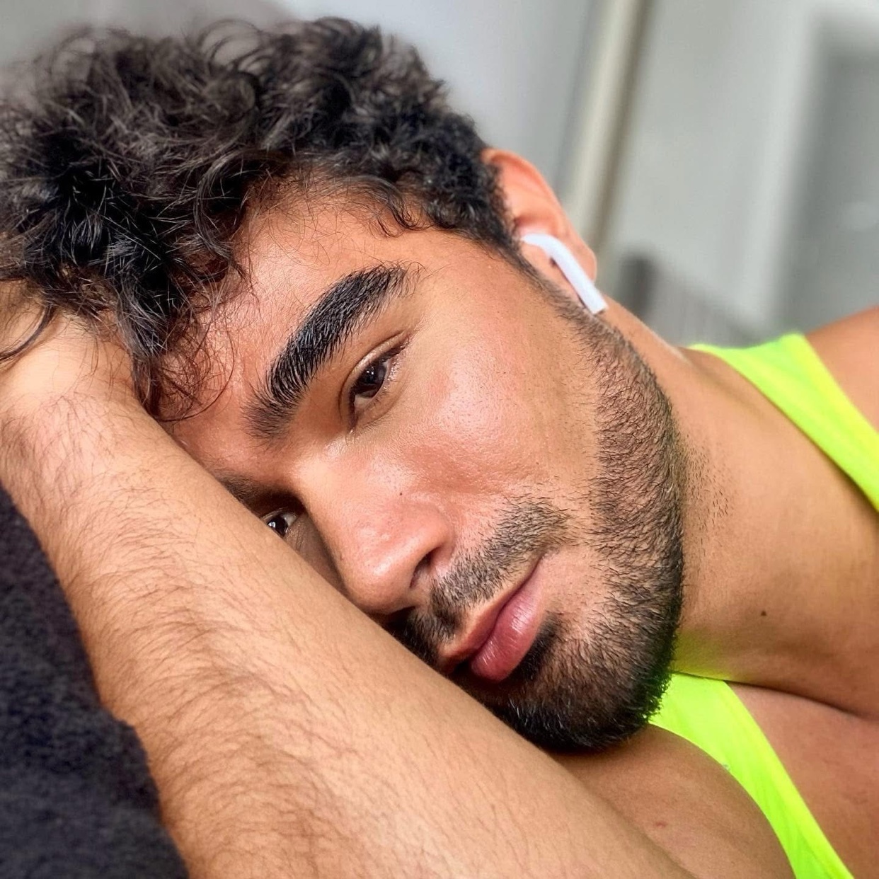 A close-up selfie of a man with curly dark hair and stubble, wearing earbuds and a neon yellow tank top. He is resting his head on his arm.