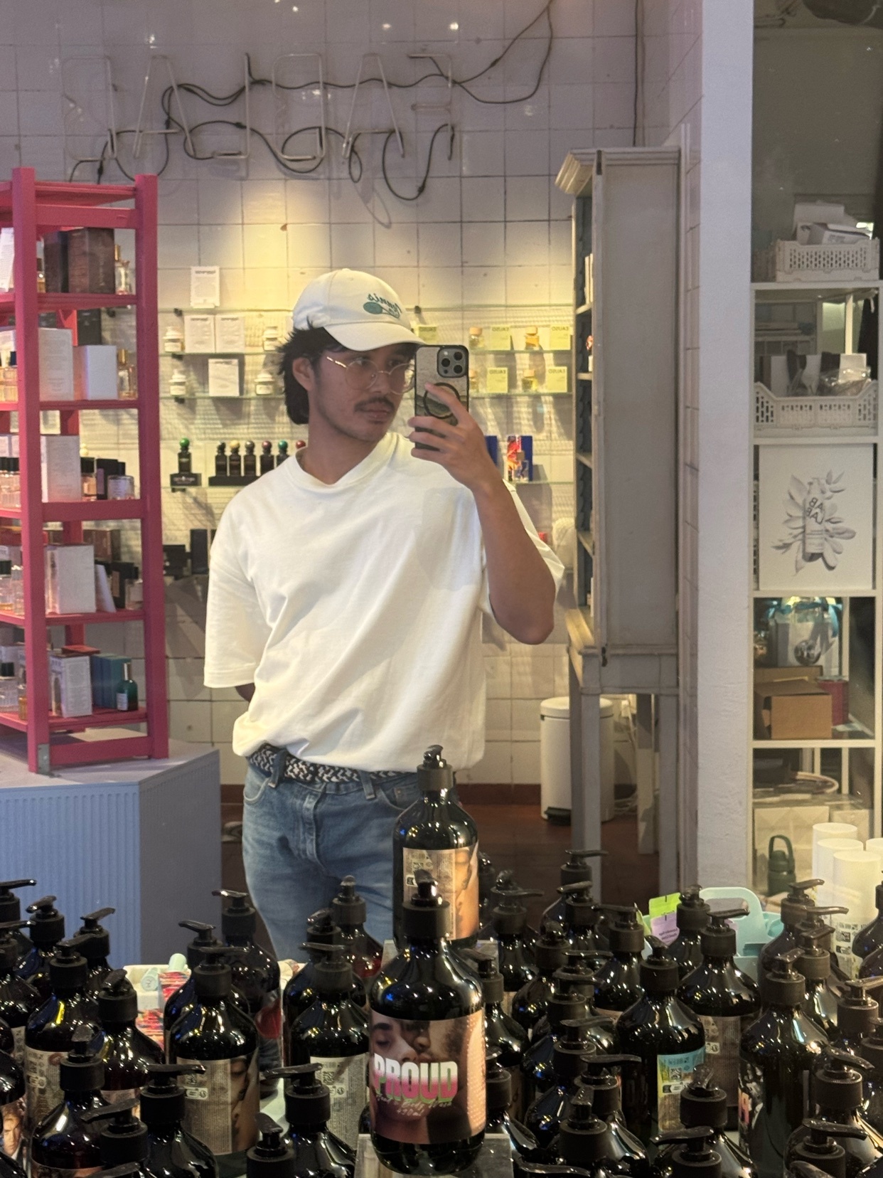 A man takes a selfie in a retail store, wearing a white t-shirt, jeans, and a baseball cap. He has stubble and a moustache, and is reflected in a mirror surrounded by bottles.