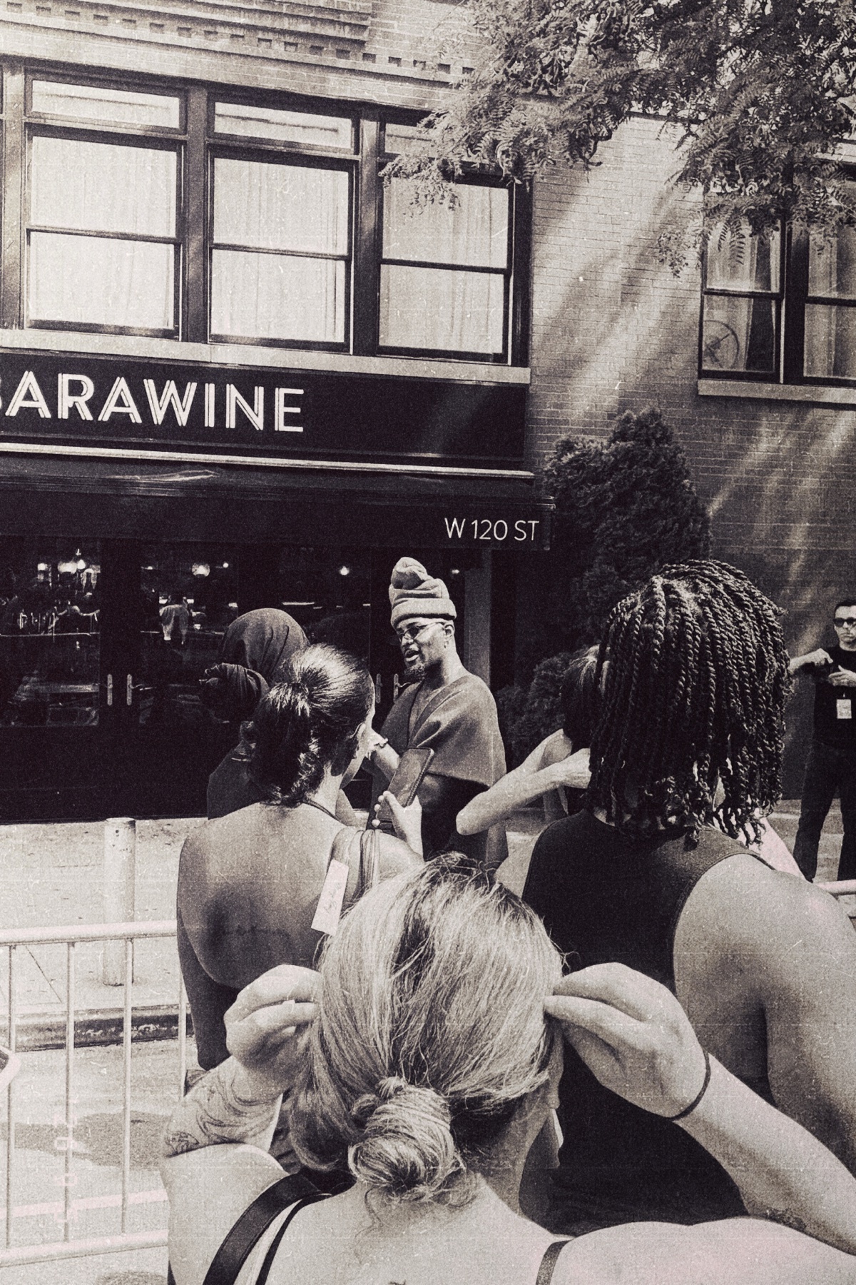 A black and white street photograph captures a group of people gathered outside a building with a sign that reads 'SARAWINE' and 'W 120 ST'. The focus is on a man in the center wearing a hat, surrounded by others whose backs are to the camera.