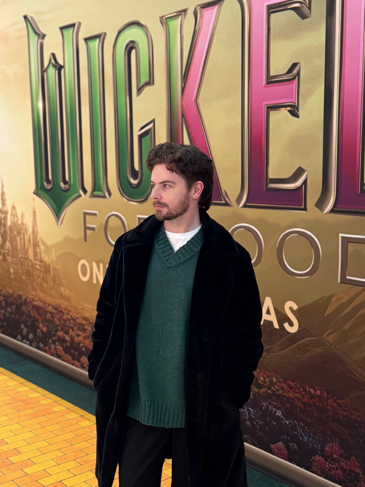 A man poses in front of a "Wicked" movie poster. He is wearing a black coat and green sweater.
