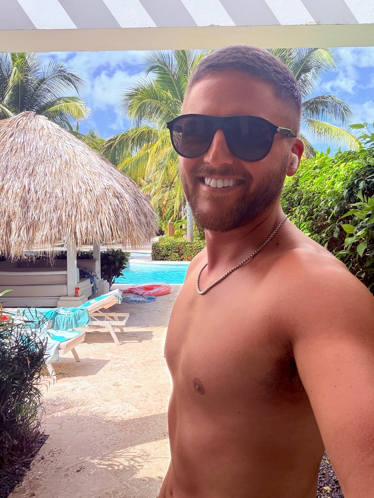 A man with short hair, a beard, and sunglasses smiles at the camera while on vacation in Punta Cana. He is shirtless, wearing a necklace and earbuds, with a pool and tropical foliage in the background.