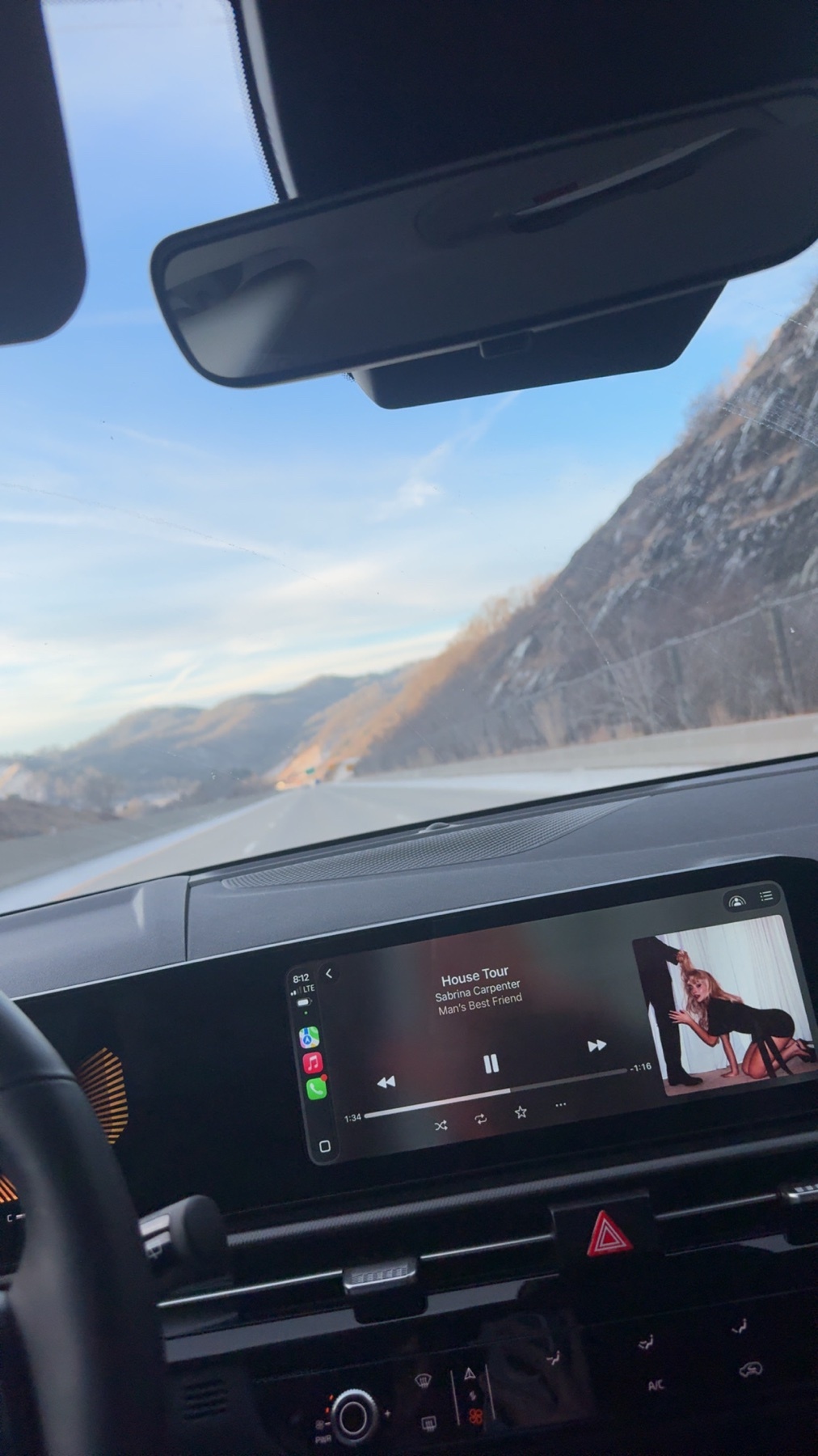 A view from inside a car, showing the dashboard and infotainment screen displaying a music video. The car is on a road with mountains in the background under a clear blue sky.