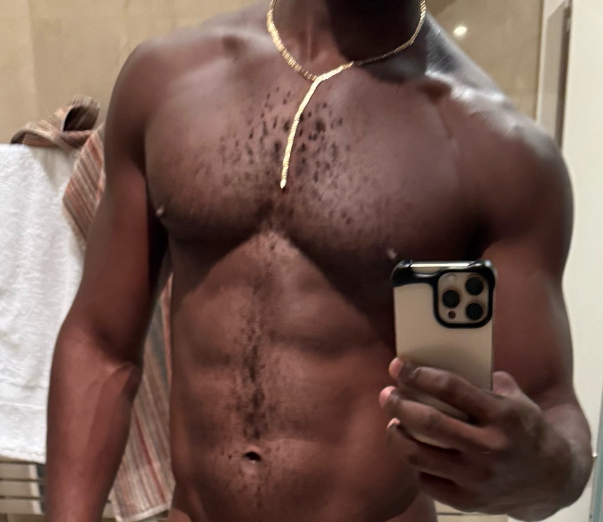 A shirtless man takes a mirror selfie, showcasing his muscular physique. He is wearing a gold necklace.