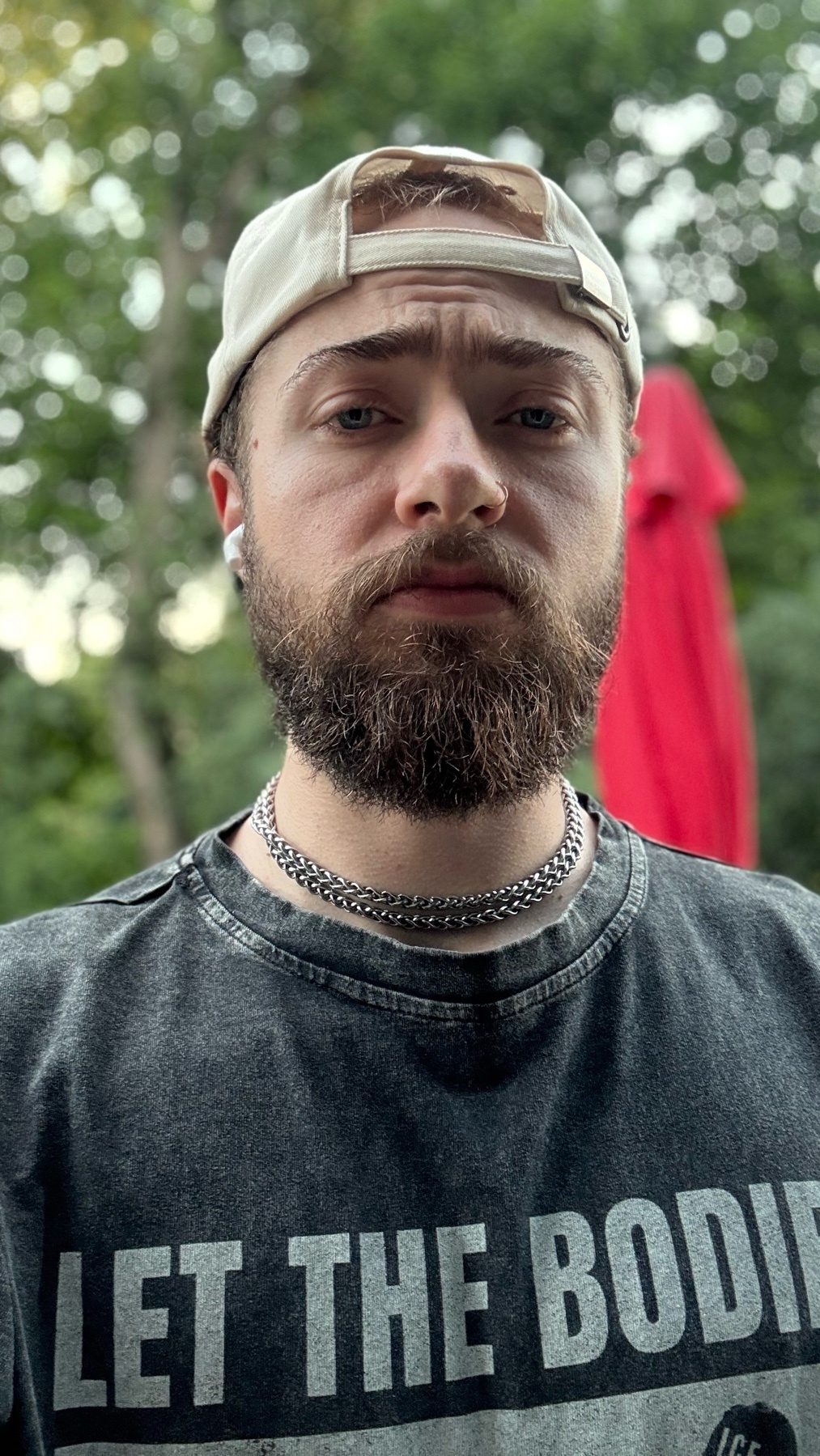 A man with a beard and a baseball cap looks directly at the camera. He is wearing a t-shirt and earbuds, with a chain necklace around his neck.