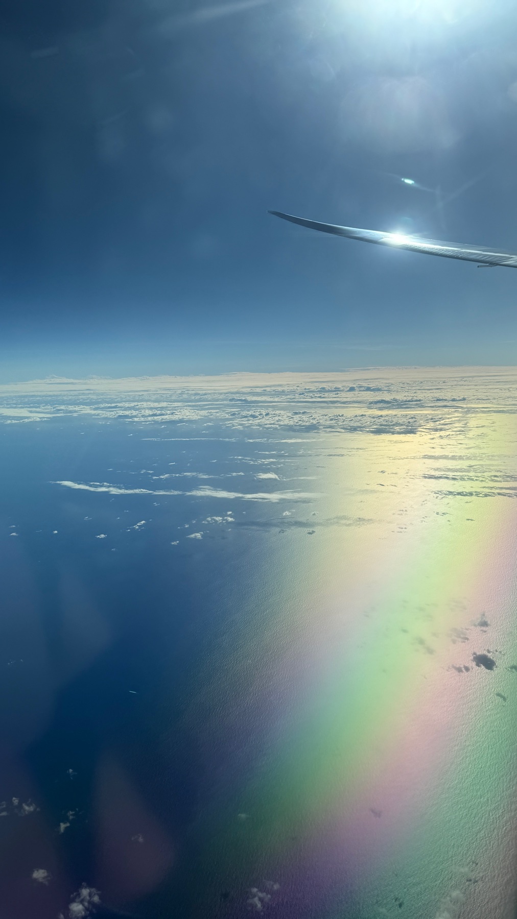 A stunning view from an airplane window shows the vast Indian Ocean below, dotted with clouds. A vibrant rainbow arcs across the water, illuminated by bright sunlight reflecting off the airplane's wing.