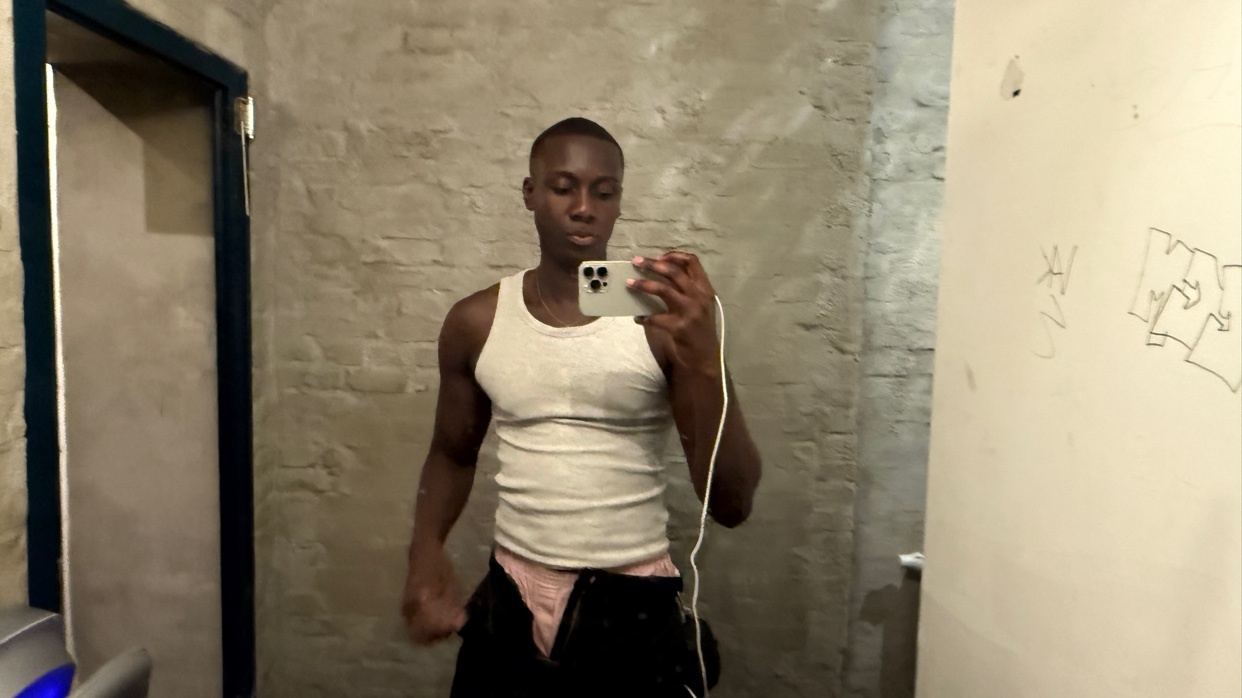 A man in a white tank top and black pants takes a mirror selfie in a bathroom.
