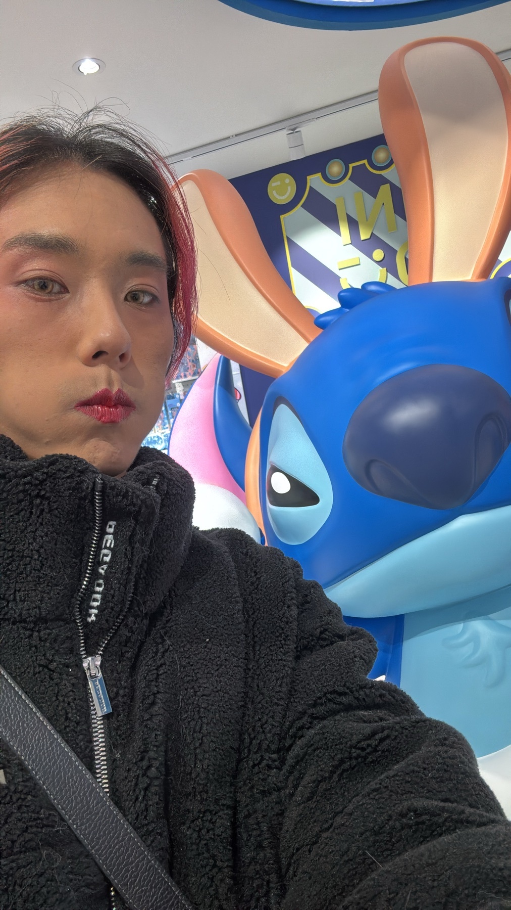 A selfie of a man with pink hair and makeup, posing with a Stitch statue. The man is wearing a black jacket.