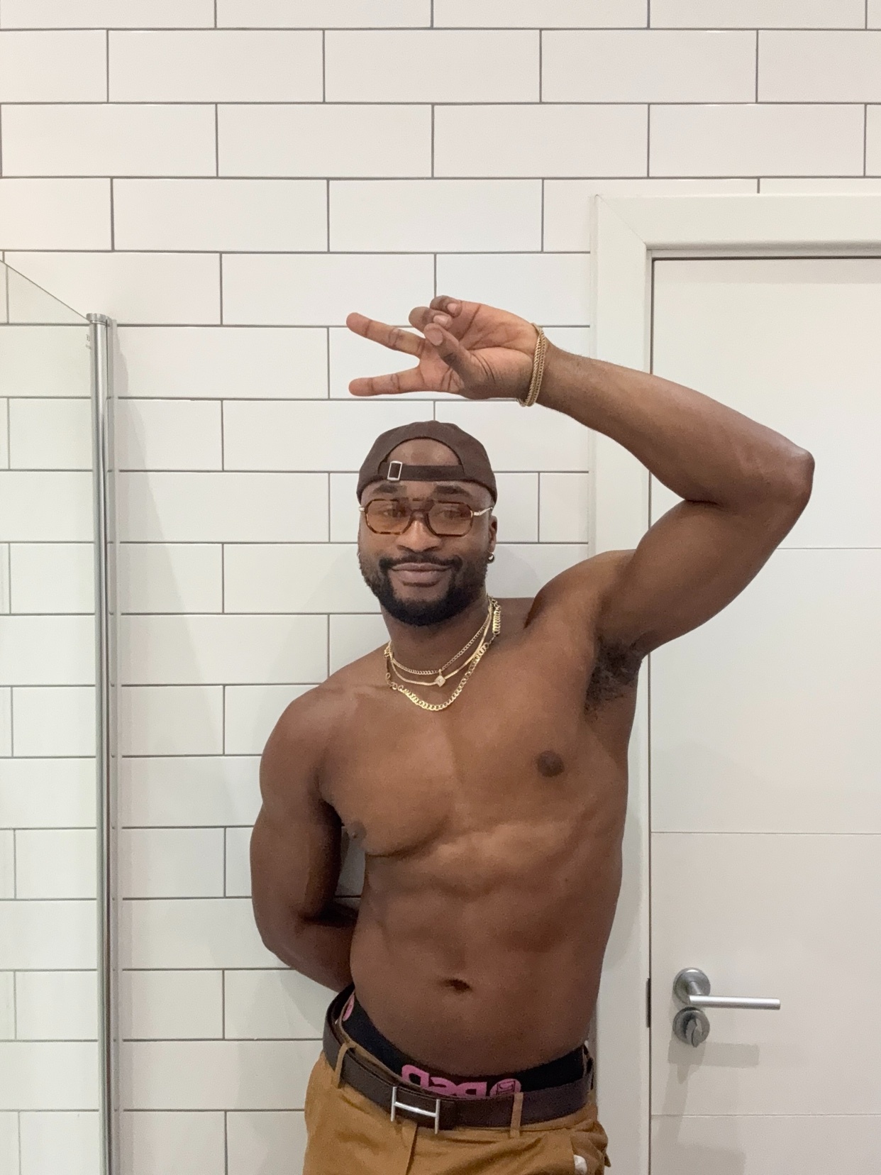 A shirtless man with muscular abs and arms poses in front of a white tiled wall, wearing glasses, a baseball cap, and gold chains. He holds up two fingers on one hand and makes a peace sign with the other.