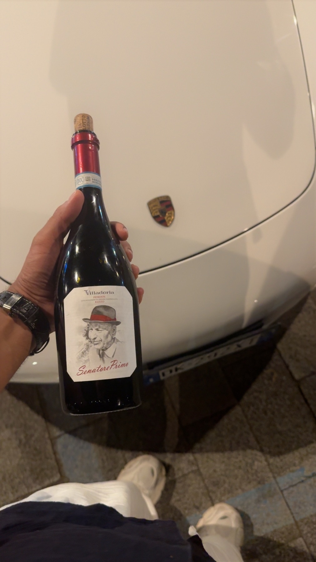 A hand holds a bottle of red wine in front of a white Porsche. The bottle has a label featuring a portrait of a man wearing a hat.