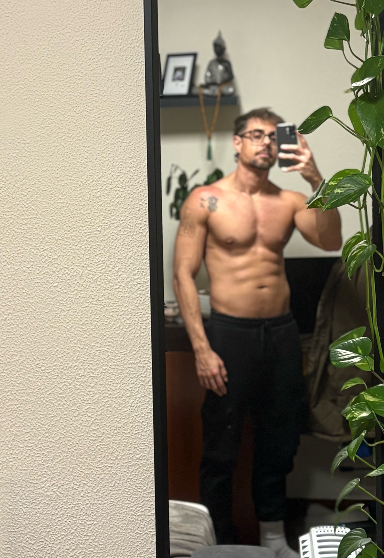 A man takes a mirror selfie, shirtless, showcasing his muscular physique. He is wearing glasses and black pants.
