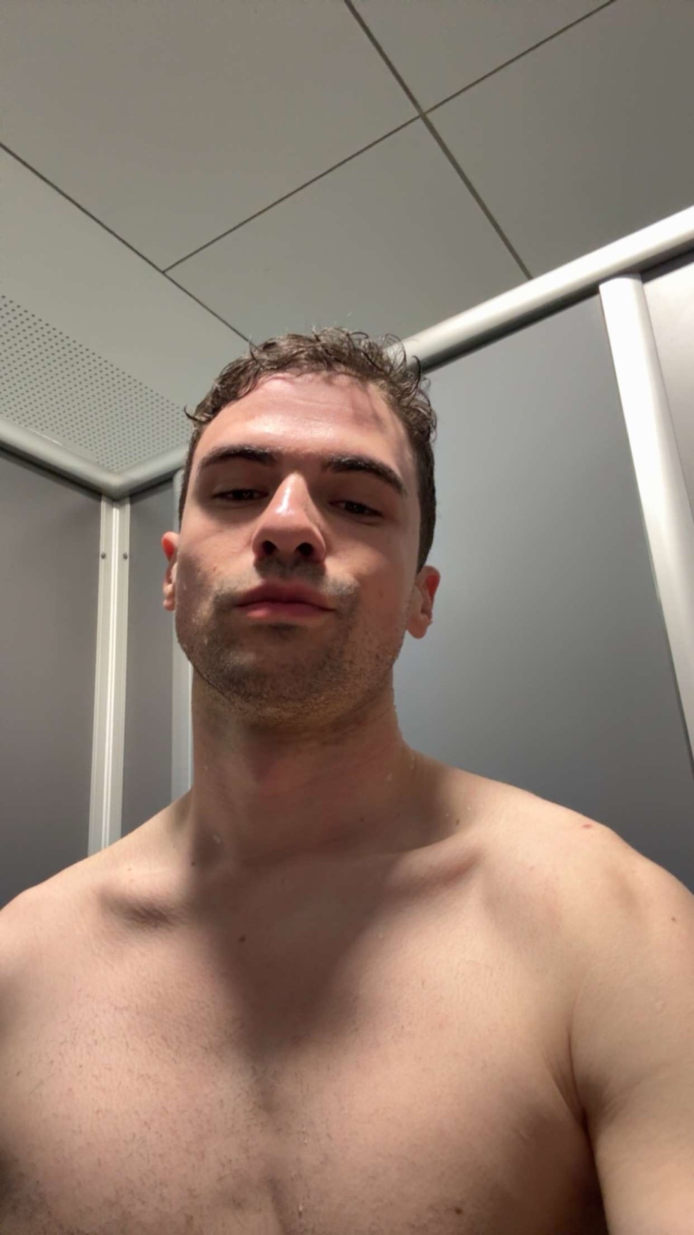A shirtless man poses for a selfie. The photo is taken in a locker room or similar setting.