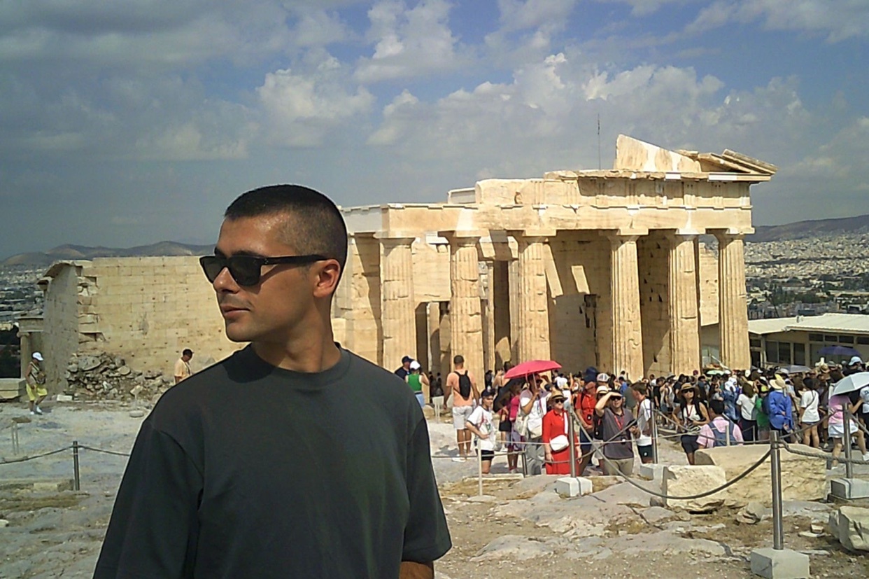 A man wearing sunglasses poses in front of the ancient ruins of the Acropolis in Athens, Greece. A crowd of tourists is visible in the background under a bright, partly cloudy sky.