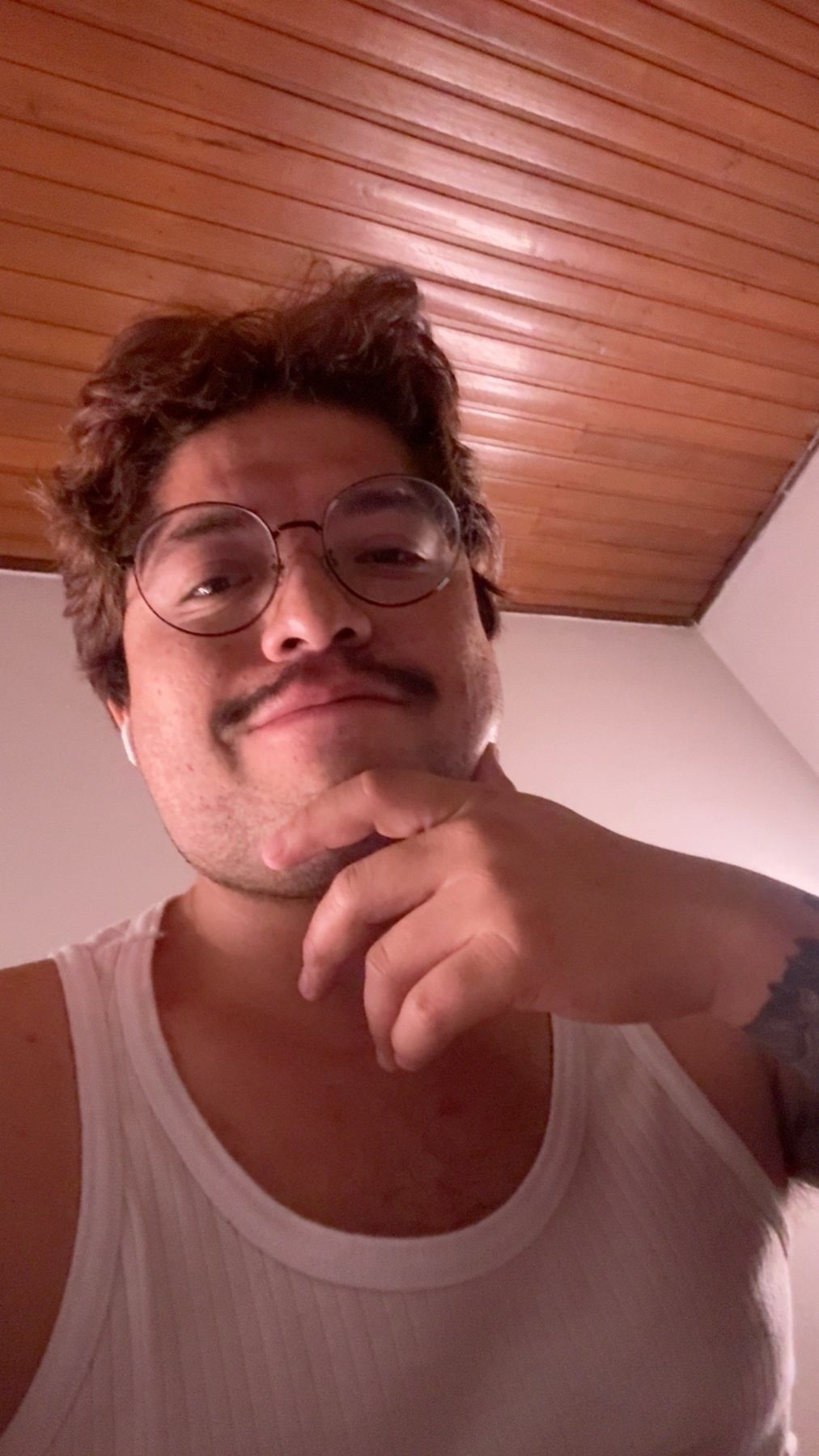 A man with a mustache and glasses poses for a selfie, wearing a white tank top and showing off a tattoo on his arm.