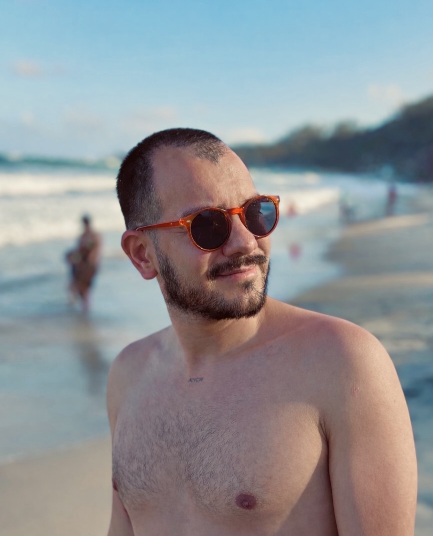 A man poses shirtless on a beach, wearing sunglasses. The ocean and sky are visible in the background.