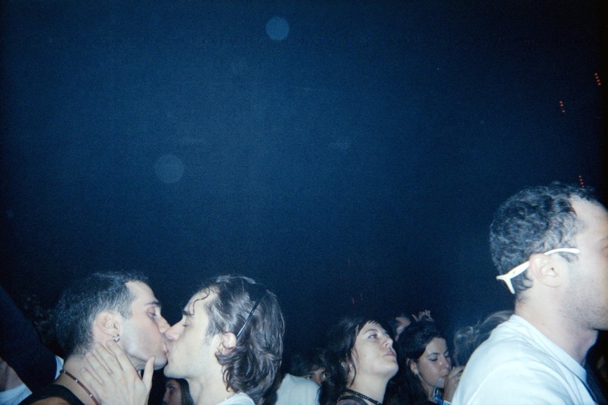 A couple shares a kiss at a rave. The photo is taken at night with a dark background.