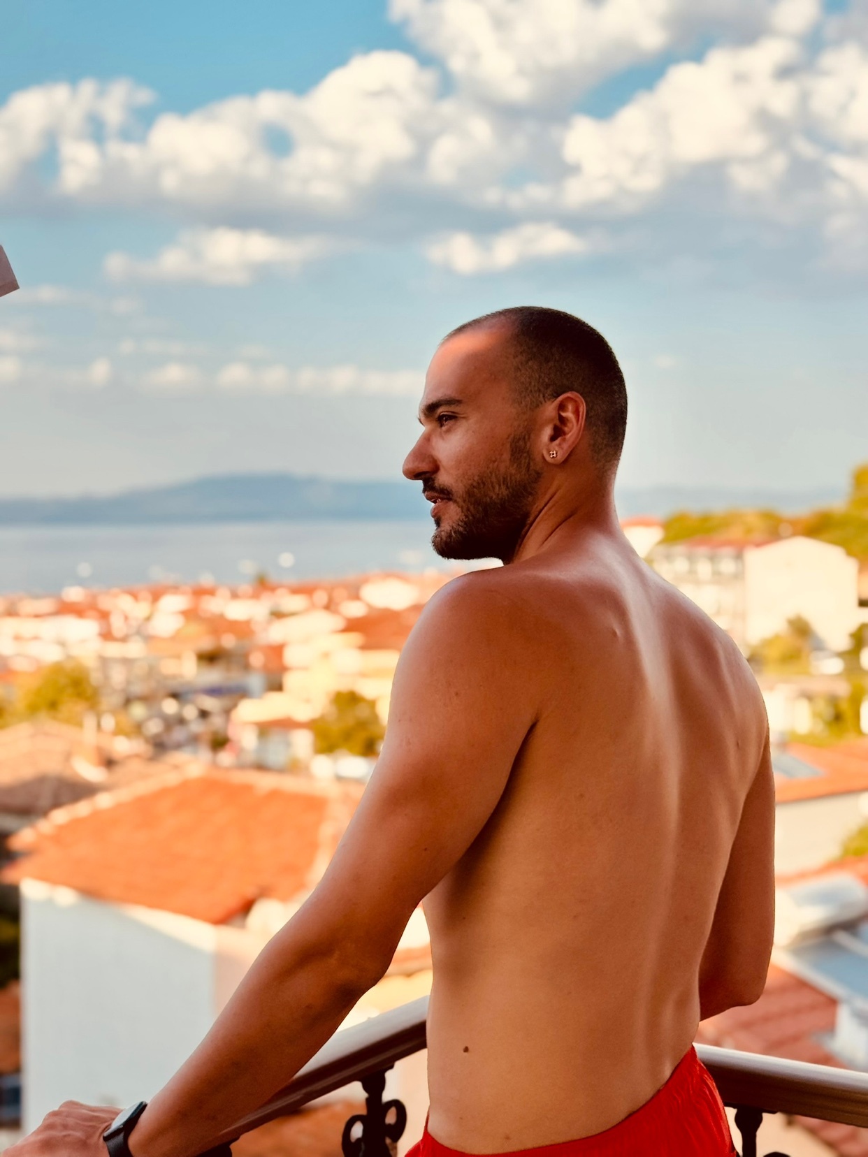 A man stands shirtless on a balcony, overlooking a cityscape and the sea. The photo captures a sunny day with a clear blue sky.