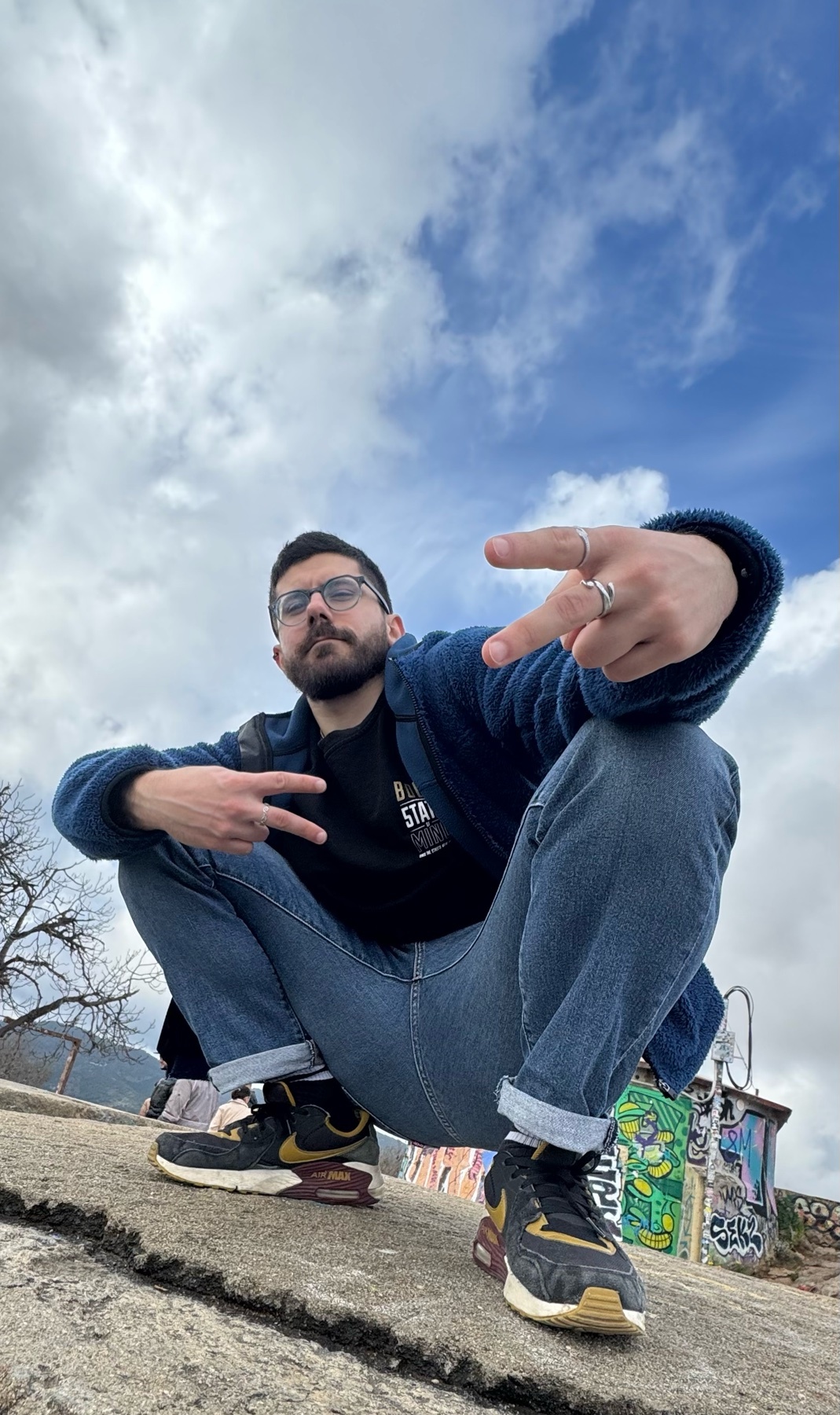 A man with glasses, a beard, and a mustache poses in jeans and sneakers against a cloudy sky. He is wearing a blue fleece jacket over a black t-shirt and holds up two peace signs with his hands.