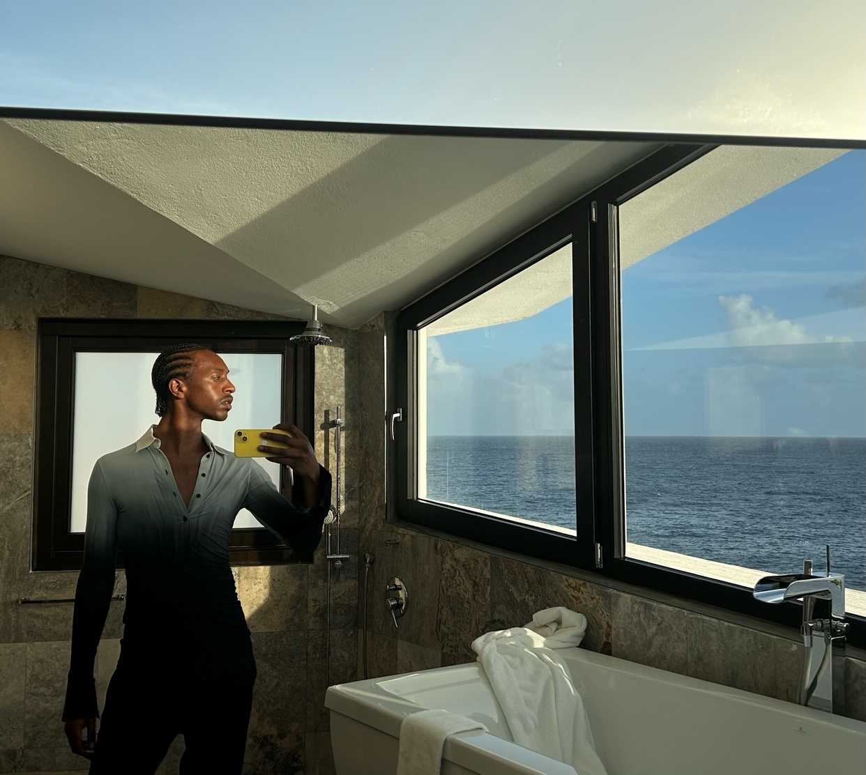 A man takes a selfie in a modern bathroom with an ocean view. He is wearing a stylish shirt and posing for the photo.