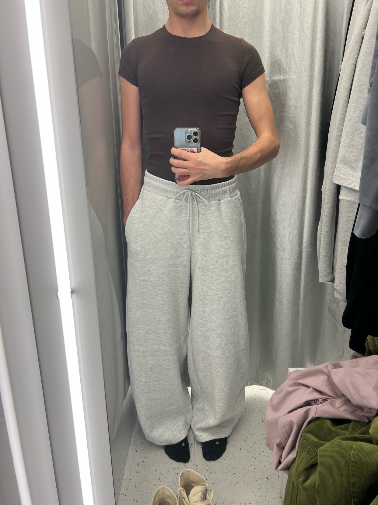A man poses in a fitting room, wearing a brown t-shirt and grey sweatpants. The photo is a mirror selfie, showcasing a modern streetwear style.