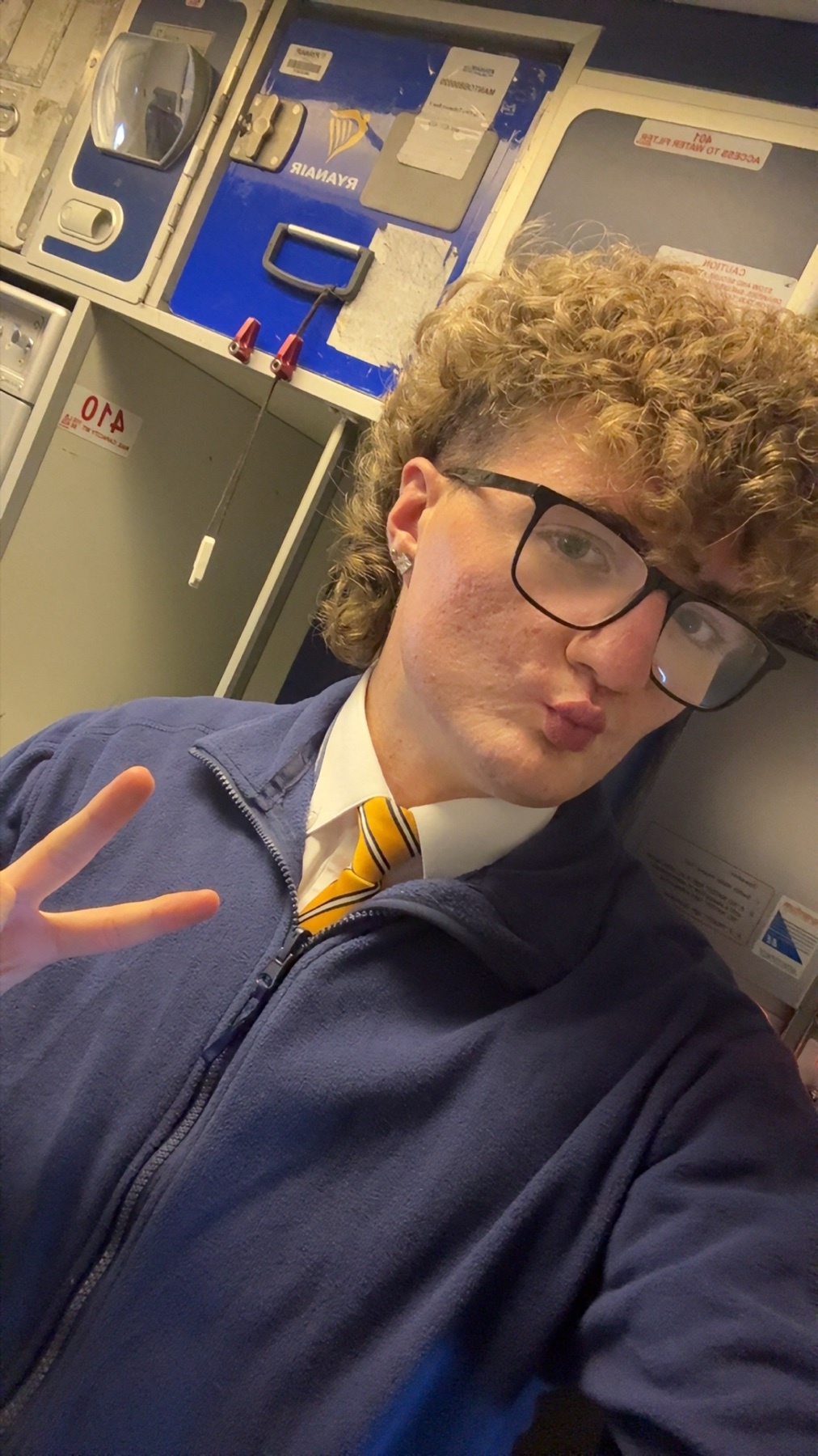 A young man with curly hair and glasses poses for a selfie in an airplane cabin, making a peace sign with his hand.
