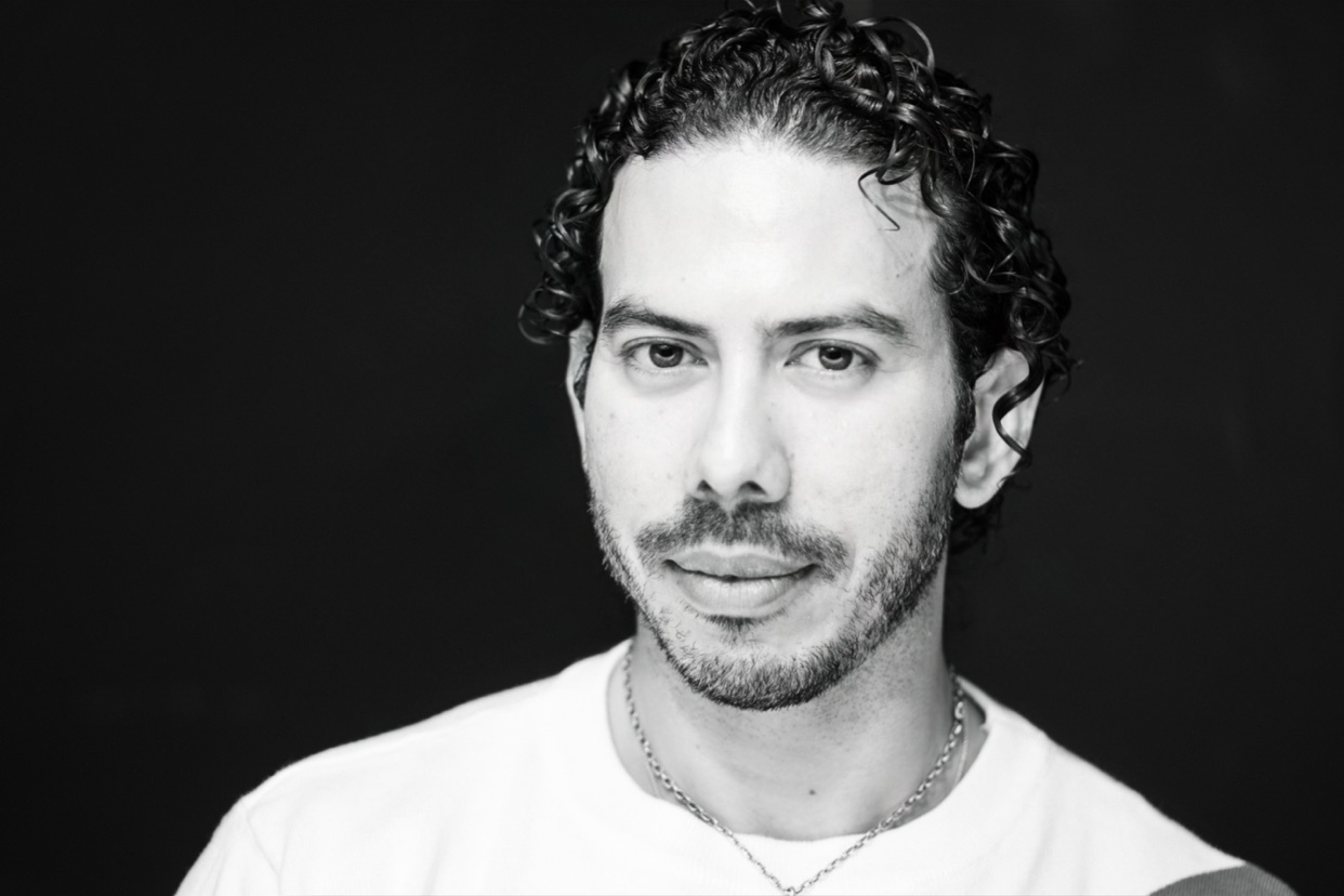 A black and white close-up portrait of a man with curly hair and stubble, he is smiling.