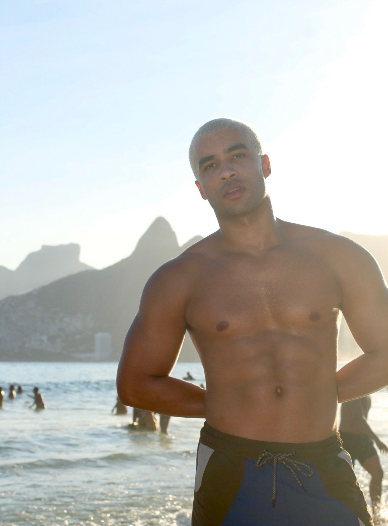 A shirtless man with a muscular physique poses on a beach in Rio de Janeiro, Brazil. The ocean and mountains are visible in the background.