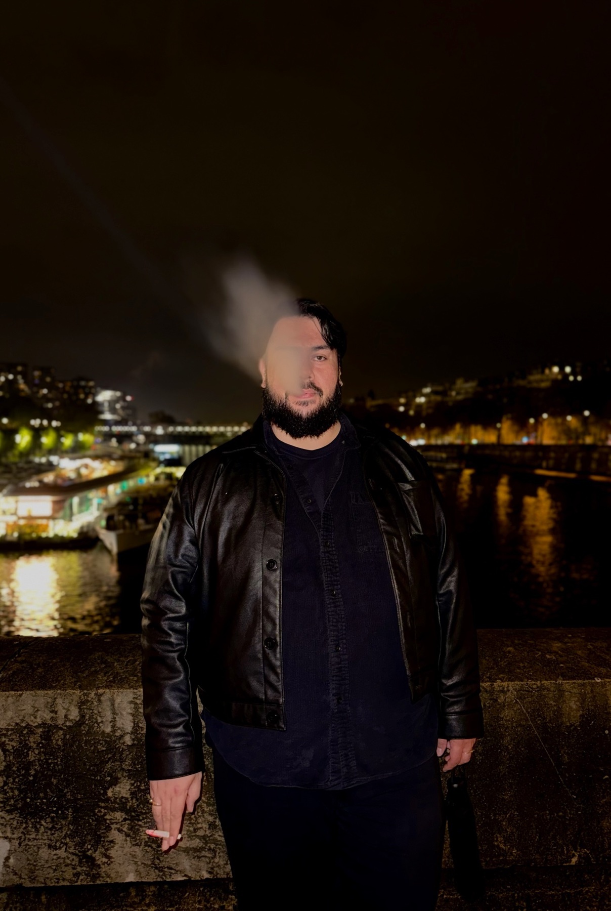 A man stands on a bridge at night, smoking and exhaling a plume of smoke. The city lights twinkle in the background, reflecting on the water.