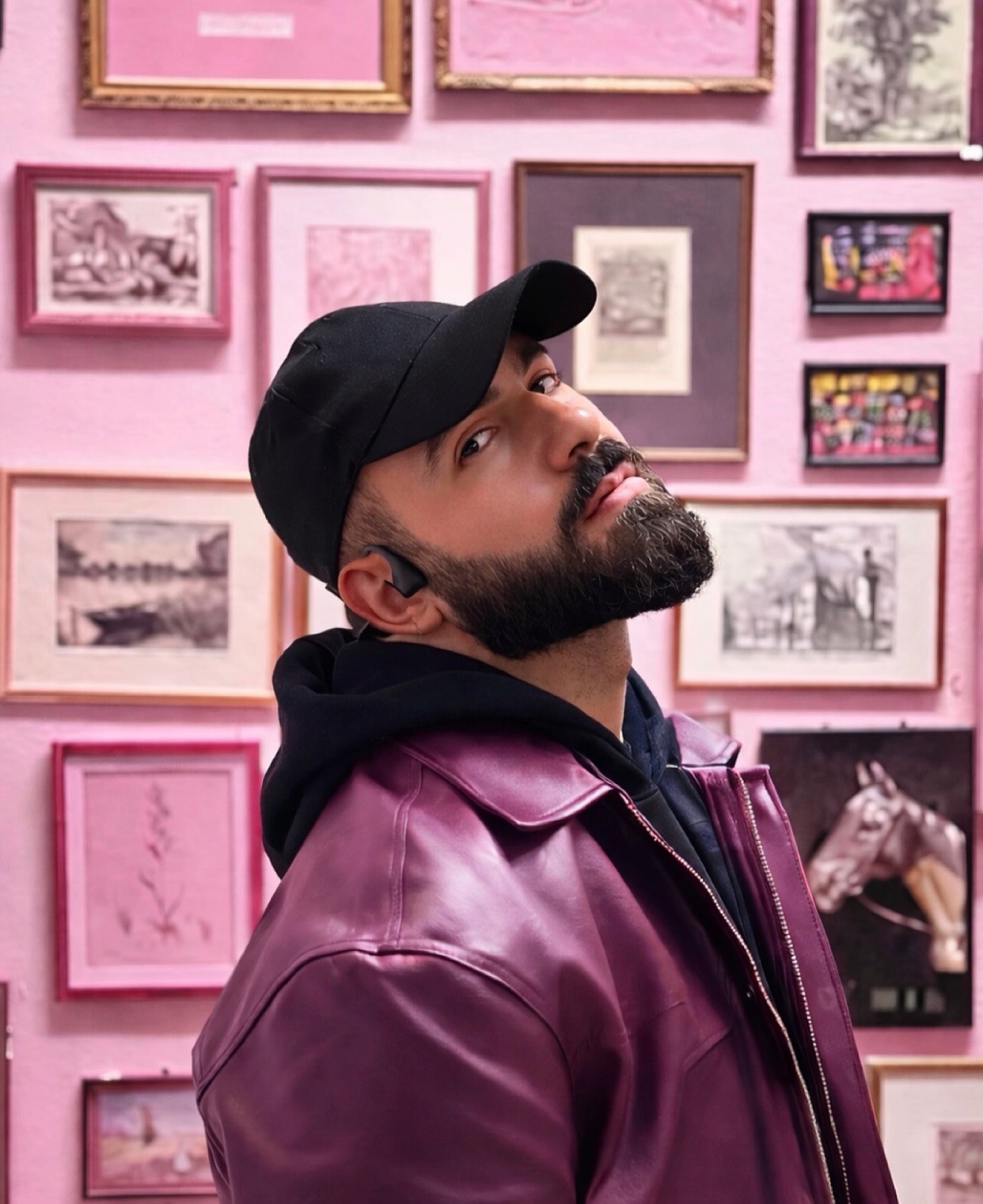A man with a beard and a black baseball cap poses against a pink wall adorned with various framed artworks. He wears a stylish purple leather jacket over a black hoodie and earbuds.