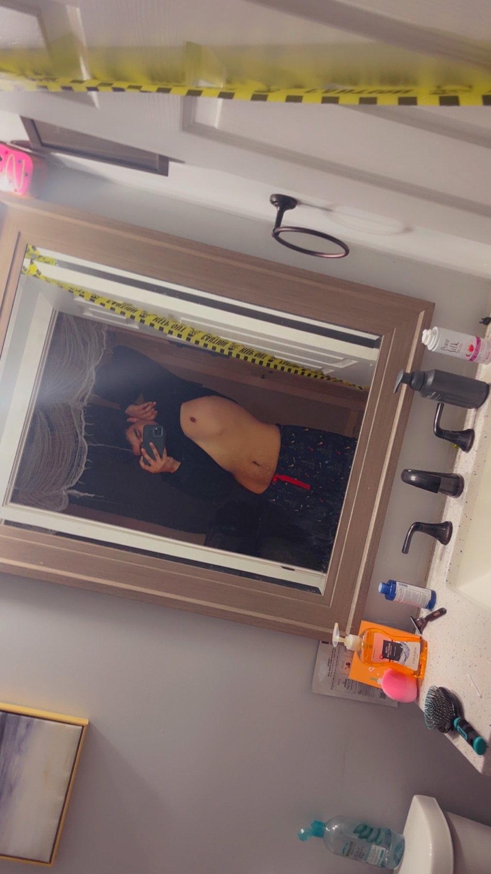 A woman takes a mirror selfie in a bathroom, showcasing her physique. She is shirtless, revealing her abs and wearing underwear.