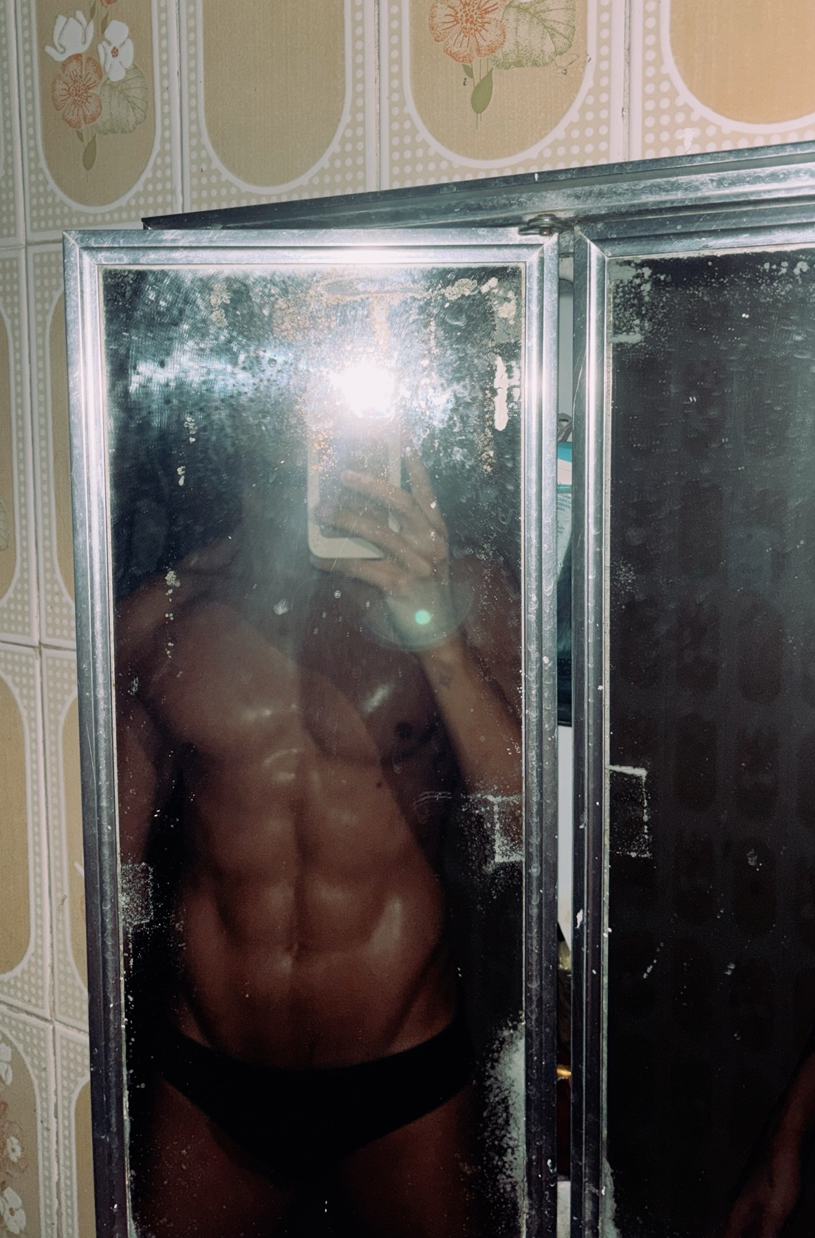 A shirtless man with visible abs takes a mirror selfie in a bathroom. He is wearing dark underwear and his body is glistening.