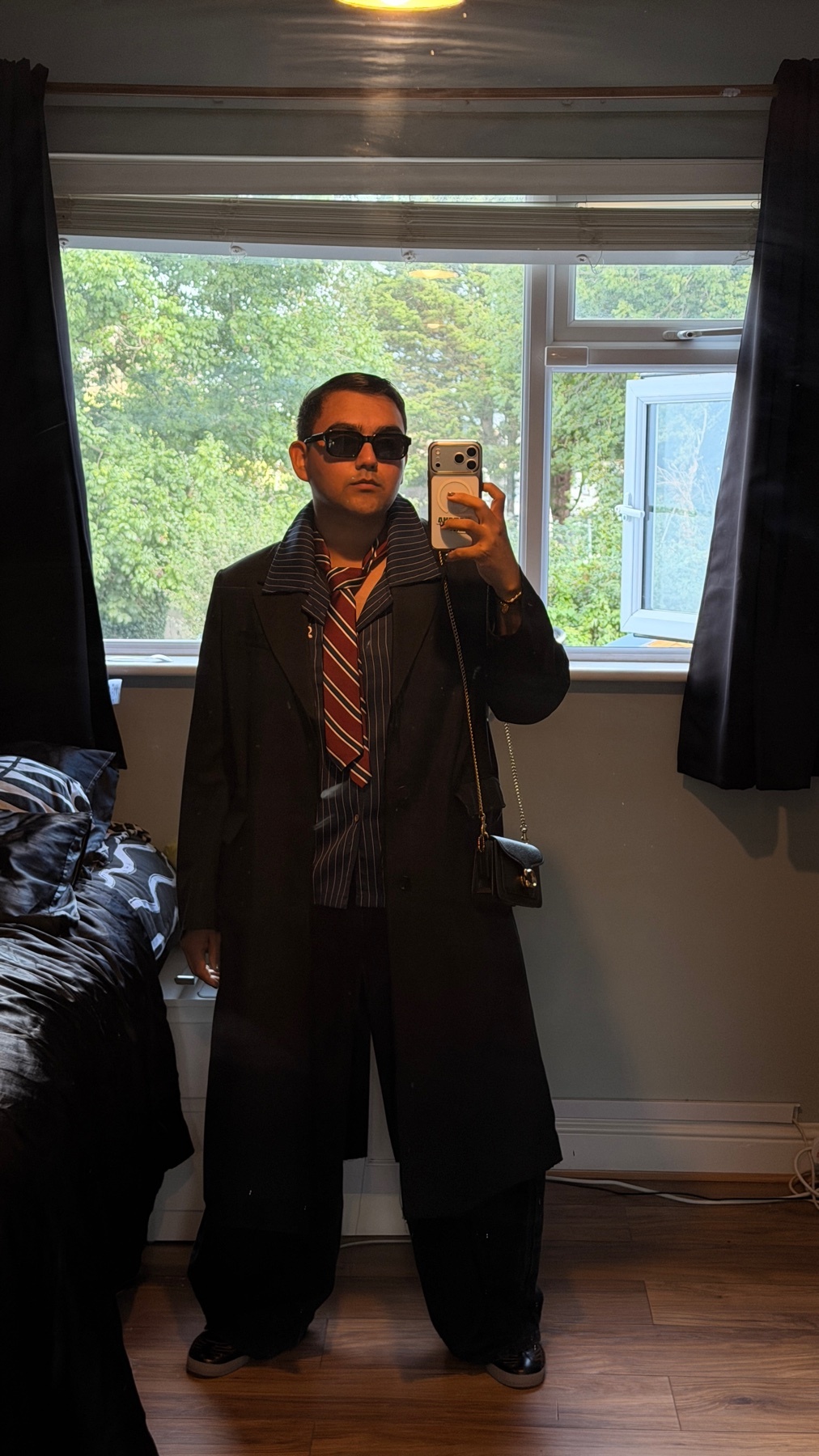 A man poses for a selfie in a dark trench coat, striped shirt, and tie. He is wearing sunglasses and wide-leg pants.