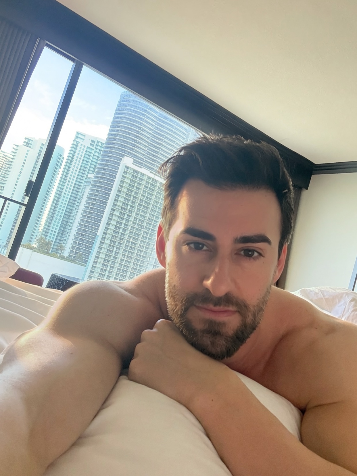 A man with stubble and dark hair lies in bed, looking at the camera. He is shirtless, with a cityscape visible through the window behind him.
