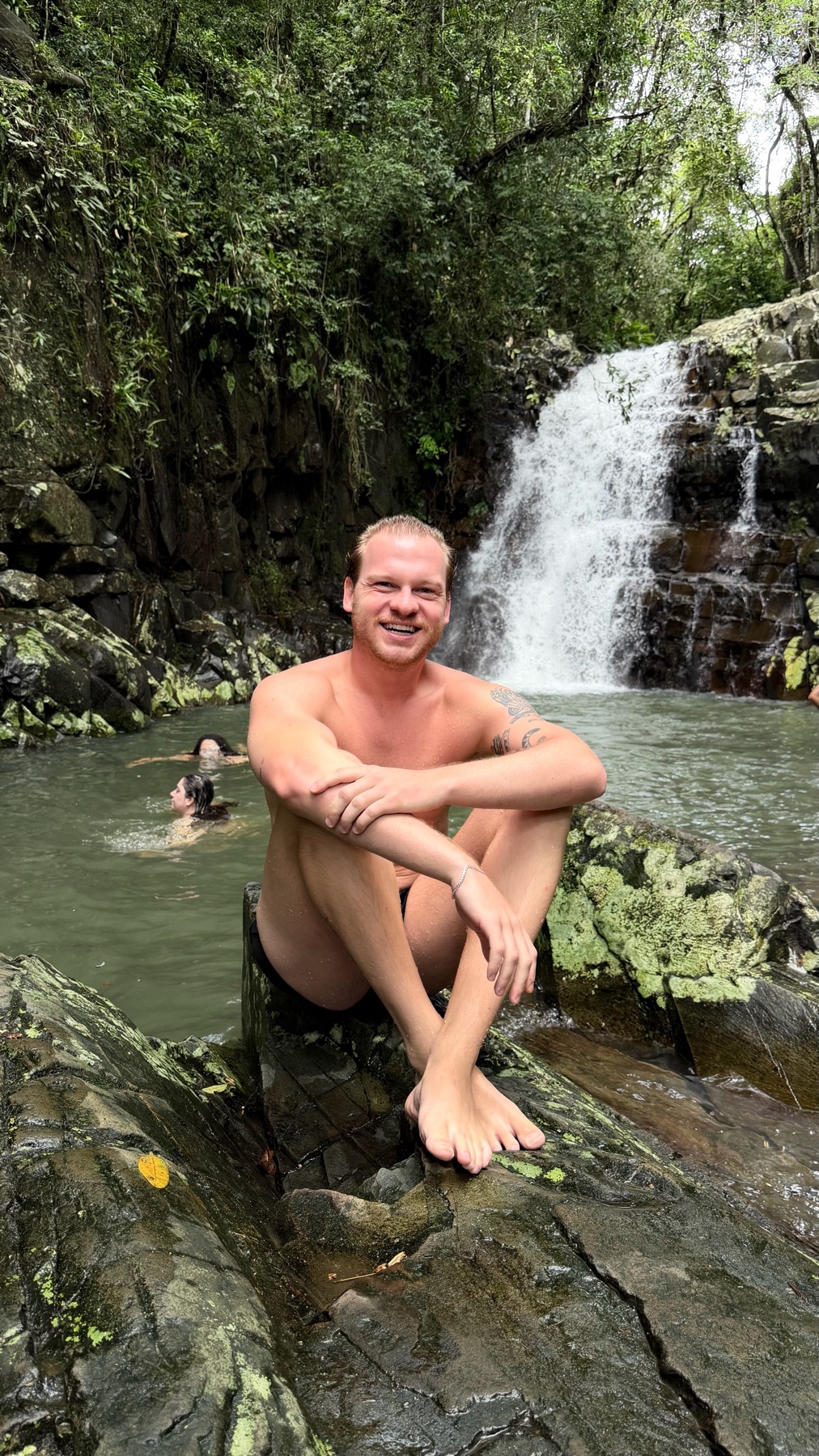 A man sits shirtless on a rock in front of a waterfall. The photo captures a moment of relaxation and connection with nature.