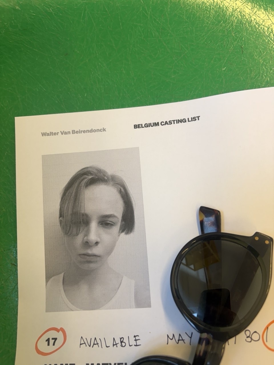 A black and white headshot of a young person with a serious expression is shown on a casting list. A pair of sunglasses rests on the paper, partially obscuring the text.