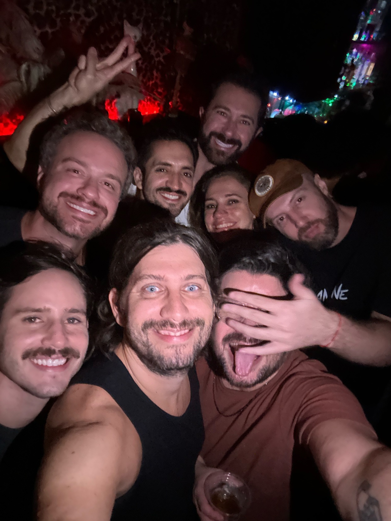 A group of seven men take a selfie in a dimly lit, possibly crowded, setting. They are all smiling and appear to be having a good time.